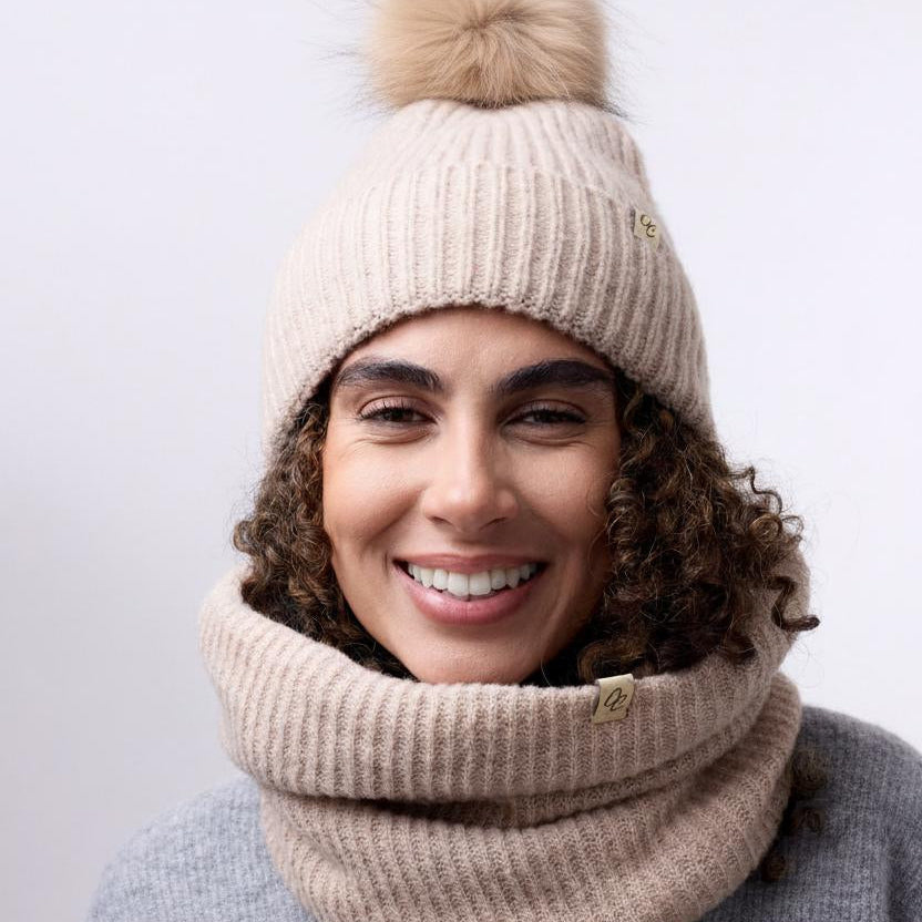 Person wearing a beige knit hat with a pom-pom and matching scarf against a white background